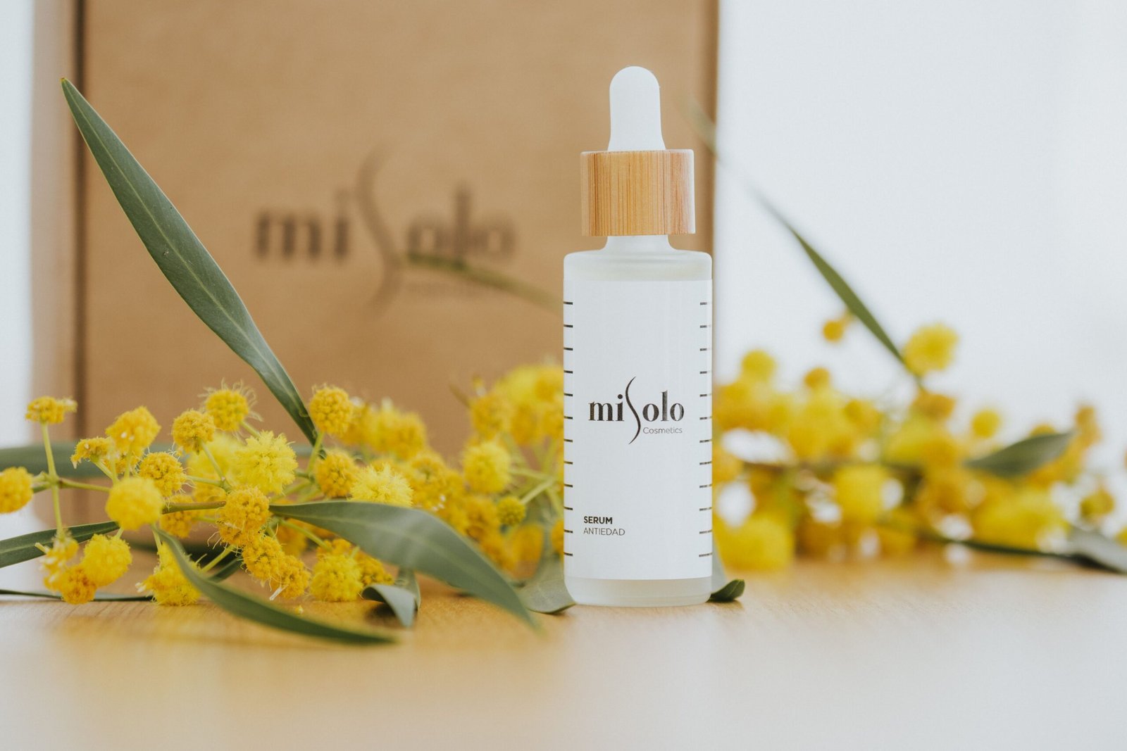 Close-up of a cosmetic serum bottle surrounded by yellow flowers on a wooden surface.